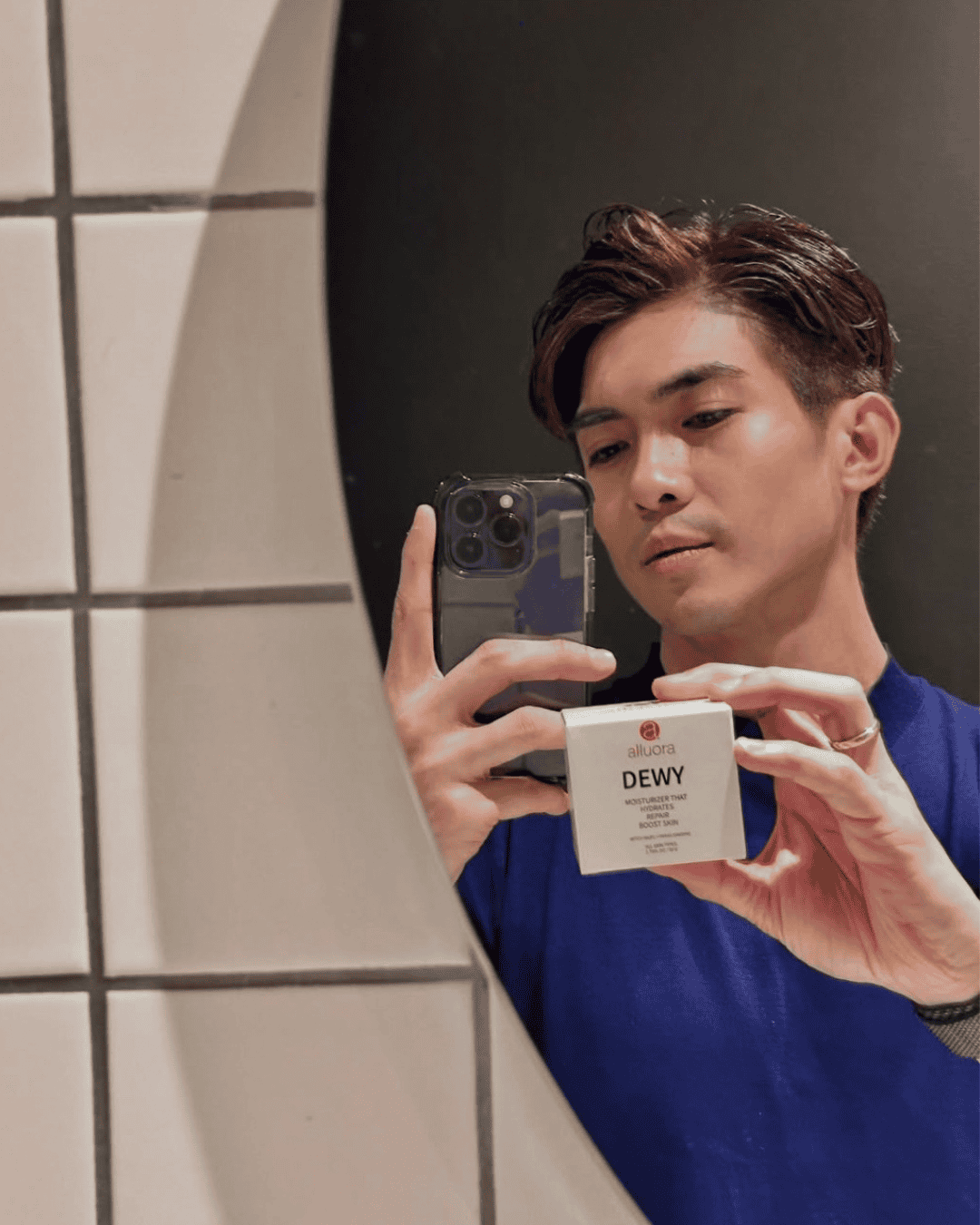 Moisturizer skincare product promotion, young man holding Alluora Dewy moisturizer, a men's skincare routine mirror selfie in bathroom.