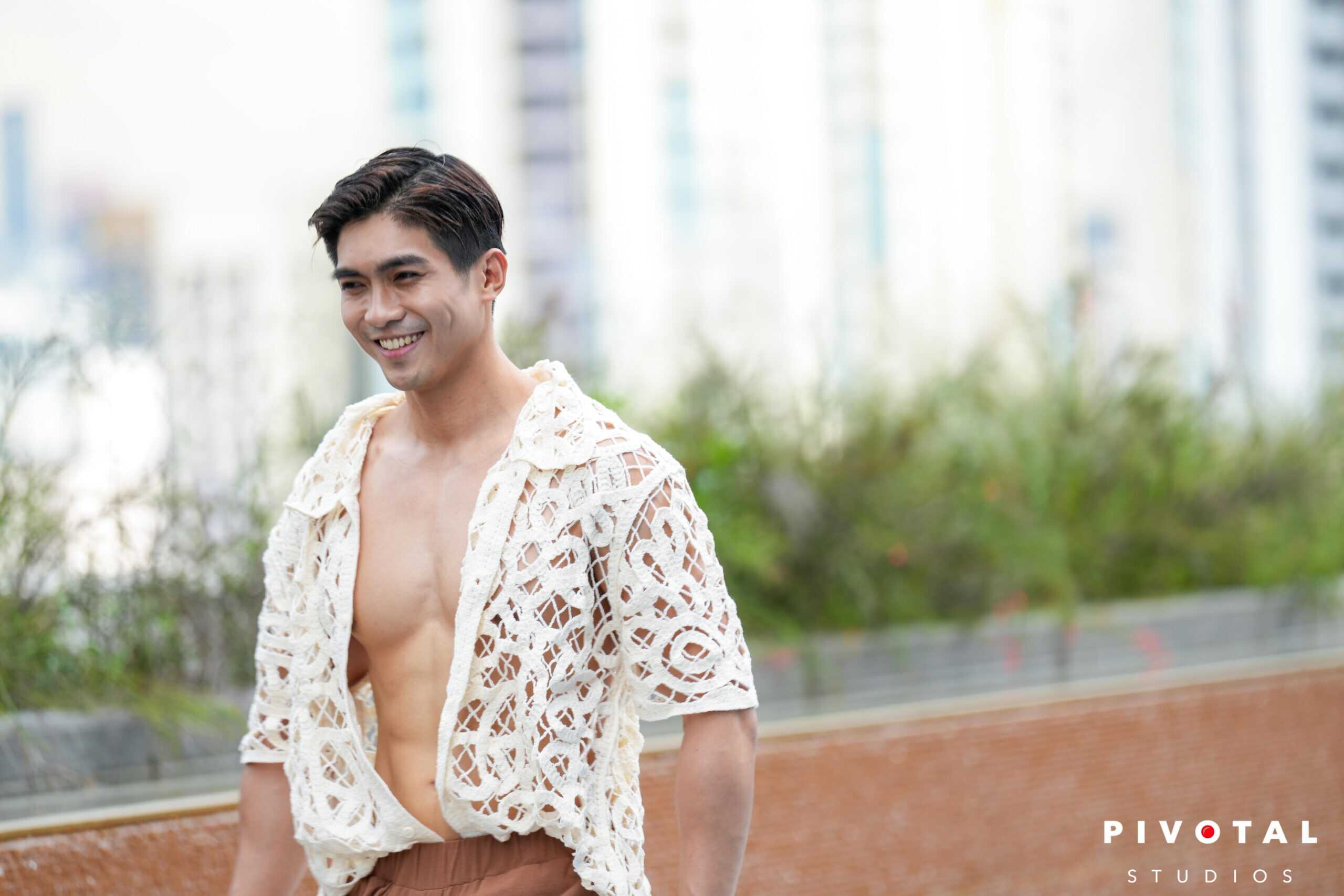 Vibrant male model wearing a stylish cream crochet cover-up and brown shorts outdoors in an urban park setting.